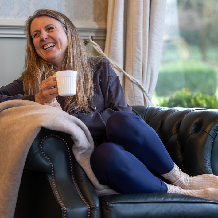 Woman sitting on a couch holding a mug, wrapped in a blanket, with a window in the background.