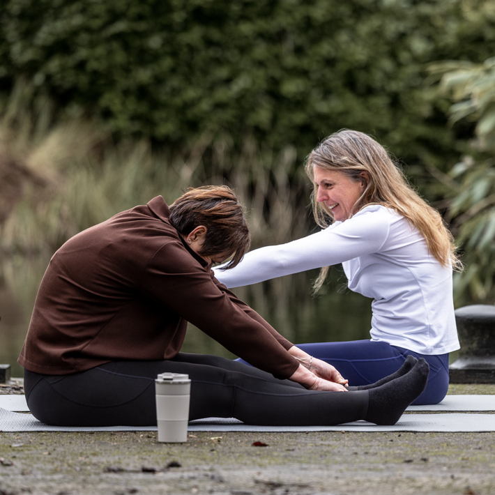Two people sitting on yoga mats outdoors with a coffee cup between them.