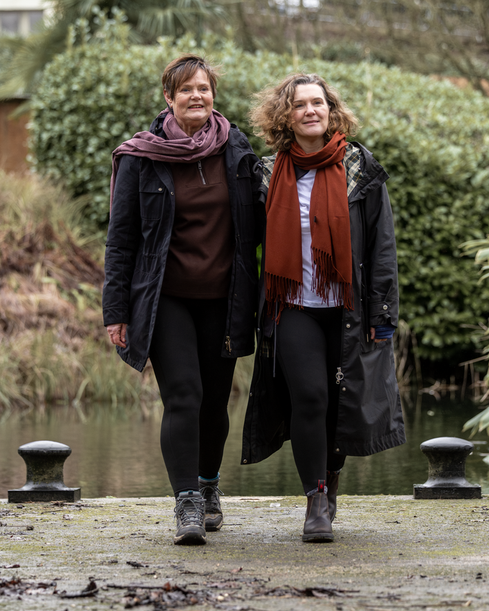 Two women walking outdoors by a pond, wearing long coats and scarves.