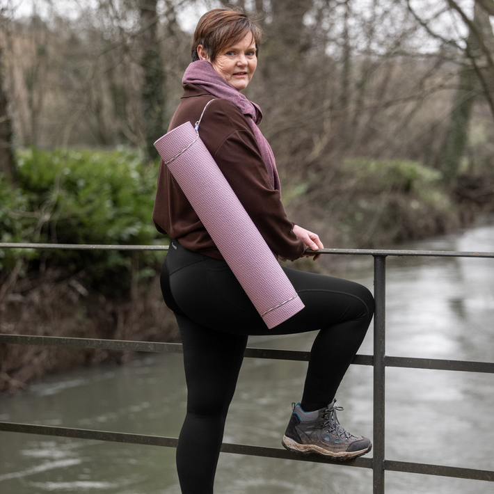 Person holding a pink yoga mat outdoors by a body of water wearing Fyness Leggings