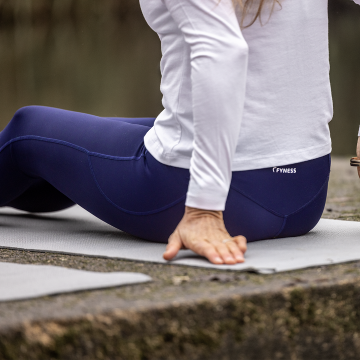 Person wearing navy leggings with a visible Fyness logo, sitting on a mat outdoors.